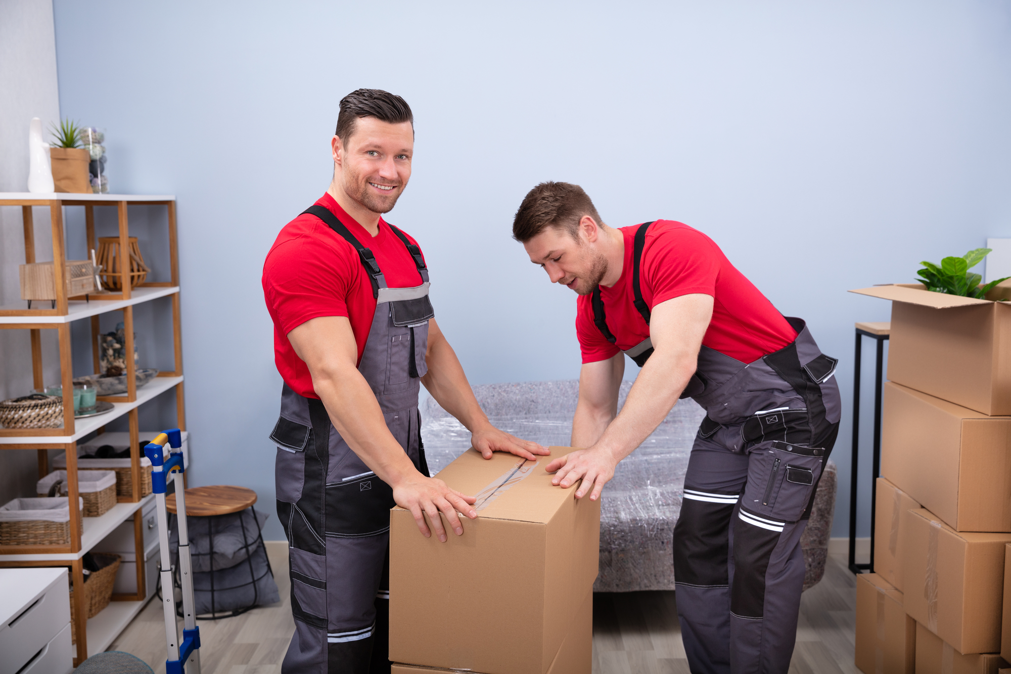 Portrait Of Two Smiling Young Male Movers In Uniform Packing The Cardboard Boxes