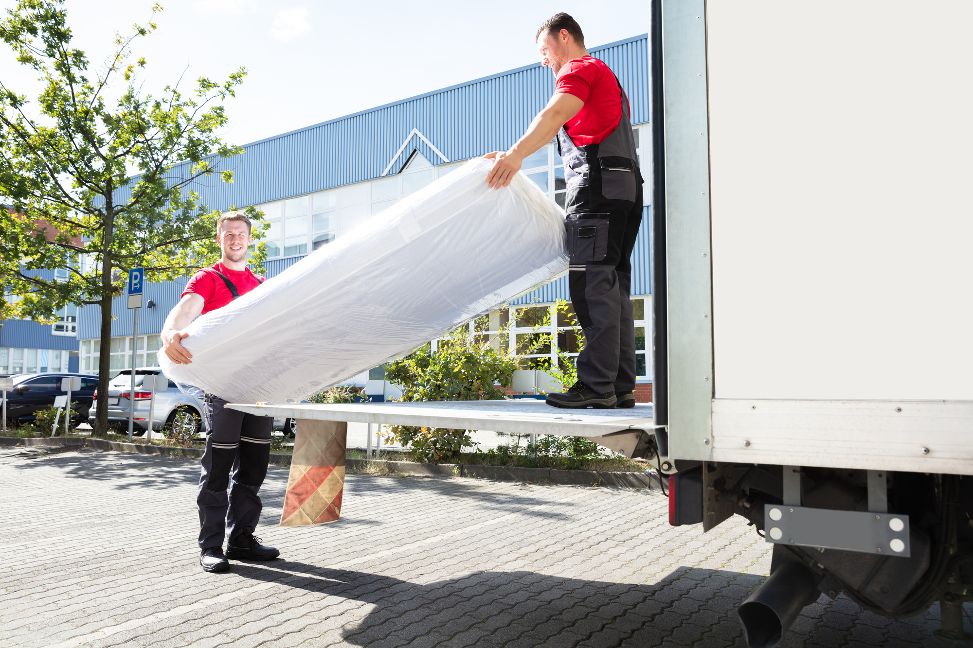 Young Men Unloading The Mattress From Truck On Street